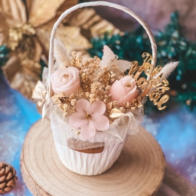 Cesta branca decorativa com flores rosa, detalhes dourados, fita de organza e mensagem Feliz Natal numa base de madeira