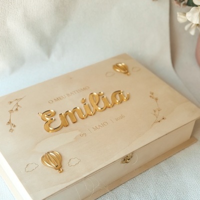 Caixa de madeira clara com texto dourado Emília para batismo e decoração de balões e flores.