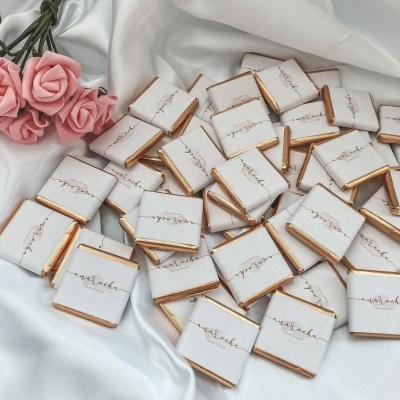 Chocolates pequenos embrulhados sobre tecido branco com ramo de rosas cor-de-rosa