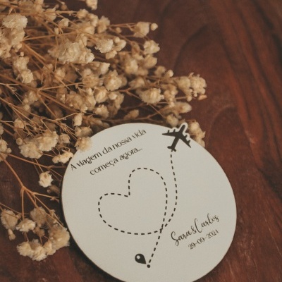 Cartão circular com texto e desenho de avião ao lado de flores secas sobre mesa de madeira