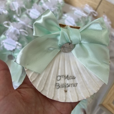 Concha de mar branca com fita verde e texto O Meu Batismo segura em mão