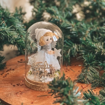 Boneca natalícia dentro de copo de vidro com 'Feliz Natal' em base de cortiça, rodeada por ramos de pinheiro