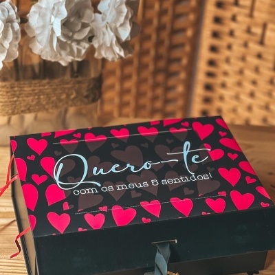 Caixa de presente preta com corações vermelhos e frase romântica colocada sobre mesa de madeira com fundo em vime.