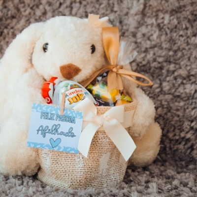 Pelúcia de coelho creme segurando saco de juta com doces e etiqueta de Feliz Páscoa