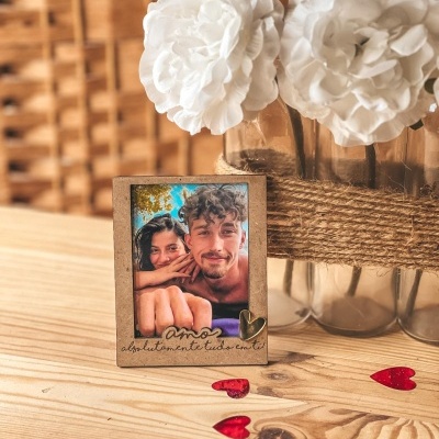 Moldura de madeira com frase romântica e foto de casal, ao lado de vaso com flores brancas e corações vermelhos sobre mesa de madeira.