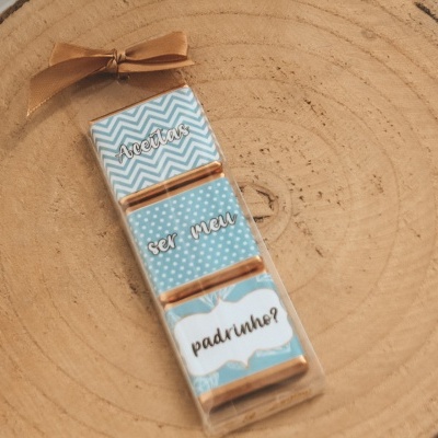 Embalagem retangular azul com texto 'Queres ser meu Padrinho?' sobre base de madeira
