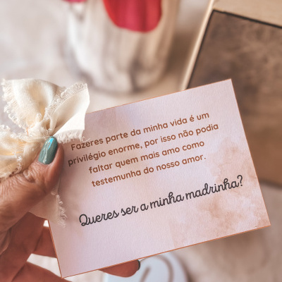 Cartão de convite em português a pedir para ser madrinha, com laço branco e mão segurando o cartão