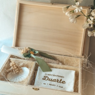 Caixa de madeira aberta com vela, concha, saco com texto de batismo e flores ao lado