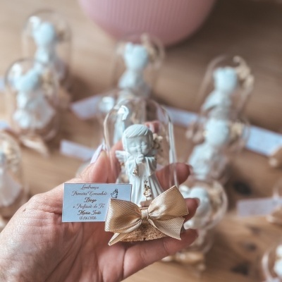 Figurinha decorativa branca com cúpula de vidro, base de cortiça, laço dourado e etiqueta com texto, várias ao fundo sobre mesa de madeira.