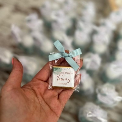 Pequeno pacote decorativo com laço azul e texto sobre batizado de Tomás em mão, pacotes semelhantes ao fundo.