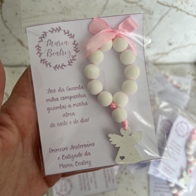 Pulseira branca com laço rosa e pendente de anjo em embalagem transparente com cartão escrito em português.