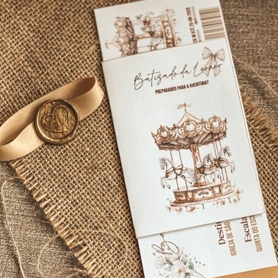 Convite de batizado com ilustração de carrossel e texto em português, sobre tecido de juta com fita bege e selo de cera dourado
