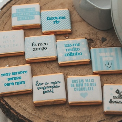Nove pequenos chocolates embalados com mensagens em português sobre uma placa de madeira.