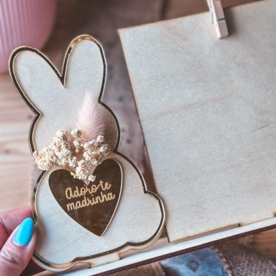 Figura decorativa em forma de coelho com coração dourado e texto, pena rosa e flores secas.