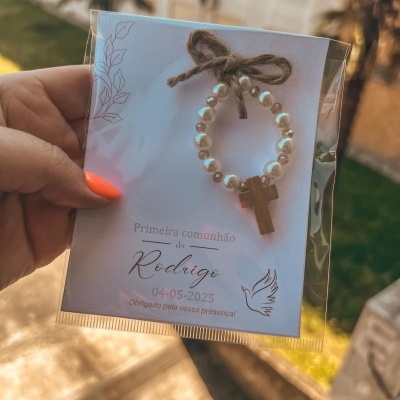 Lembrança de primeira comunhão com pulseira de contas brancas e cruz de madeira em cartão branco.