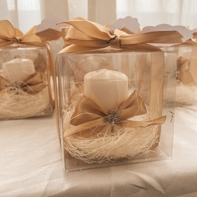 Vela branca decorativa numa caixa transparente com fita castanha e cruz metálica.
