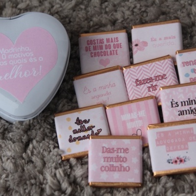 Lata de metal em forma de coração rosa claro com frases motivacionais e chocolates pequenos embrulhados em papel rosa claro e branco sobre tapete cinza.