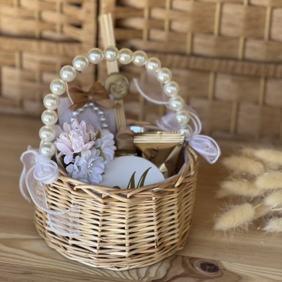 Cesto de vime com alça de pérolas, decoração em flores lilás e itens embalados, numa mesa de madeira clara