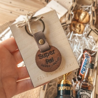 Chaveiro de madeira e pele com gravação e cartão de felicitação para o Dia do Pai, com caixa de presente ao fundo.