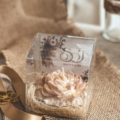 Vela decorativa em forma de flor creme dourada numa caixa transparente com etiqueta floral