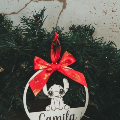 Enfeite de Natal redondo branco com personagem cartoon e nome Camila, fita vermelha com flocos de neve, entre ramos de pinheiro artificial.