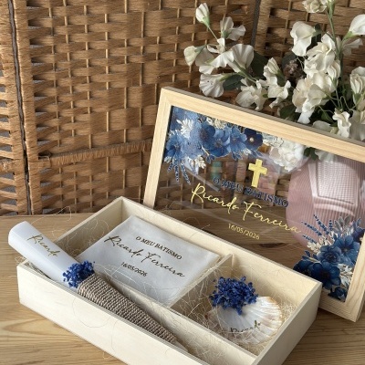 Conjunto de batismo com vela, caixa de madeira e tampa em vidro decorada com texto e flores artificiais brancas.