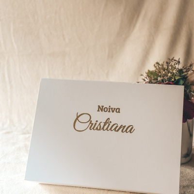 Caixa branca com texto Noiva Cristiana em dourado sobre tecido bege e flores ao fundo