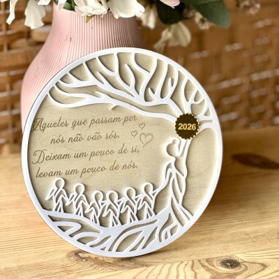 Quadro decorativo redondo com árvore e figuras humanas e texto em português, sobre mesa com vaso rosa e flores artificiais