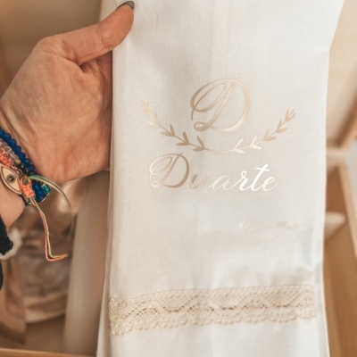 Toalha branca com renda bege e texto dourado 'D Duarte' segurada por mão com pulseiras