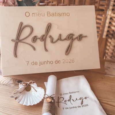 Conjunto para batismo com placa em madeira clara, vela branca, concha decorativa e pano com texto.