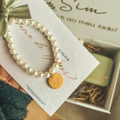 Pulseira de pérolas falsas com pendente dourado em cartolina branca junto a caixa branca com palha