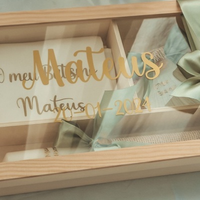 Caixa de madeira com tampa de vidro e laço verde clara contendo lembranças para bebé com texto dourado na tampa.