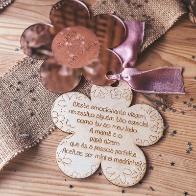 Pendural em forma de flor de madeira clara gravada com texto e pendural metálico em cobre com fita rosa sobre mesa de madeira com tecido rústico e estrelas prateadas