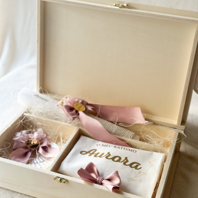 Caixa de madeira clara com acessórios para batismo incluindo vela decorada, laços rosa e saco branco com texto dourado