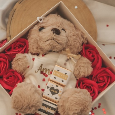Urso de pelúcia castanho claro com t-shirt branca dentro de caixa branca rodeado de flores vermelhas em fundo bege.