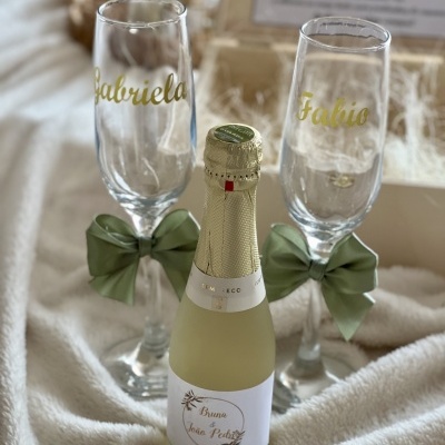 Conjunto de flautas de vidro com nomes dourados, laços verdes e mini garrafa de espumante com rótulo personalizado sobre tecido creme.