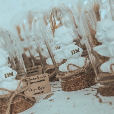 Pequenas figuras brancas dentro de redomas de vidro com base de cortiça, amarradas com cordão e etiqueta agrupadas sobre mesa