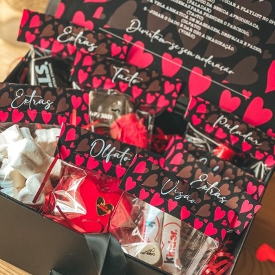 Caixa presente preta com sacos transparentes com etiquetas de corações vermelhos e castanhos e doces variados no interior.