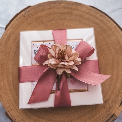 Caixa de presente branca com laço rosa e flor, em base de madeira redonda.
