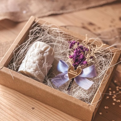 Caixa de presente com vela branca em relevo e ramo de flores secas com laço lilás e plaquinha em forma de coração.