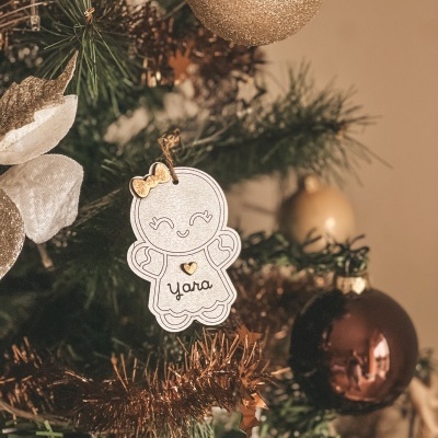 Enfeite de Natal com boneco de papel branco e dourado com nome Yara pendurado numa árvore decorada.