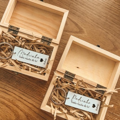Duas caixas de madeira com etiquetas 'Madrinha Gosto muito de ti' e 'Padrinho Gosto muito de ti' e argolas metálicas, sobre mesa de madeira.