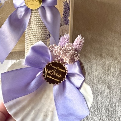 Concha branca decorada com laço lilás, flores secas e etiqueta dourada