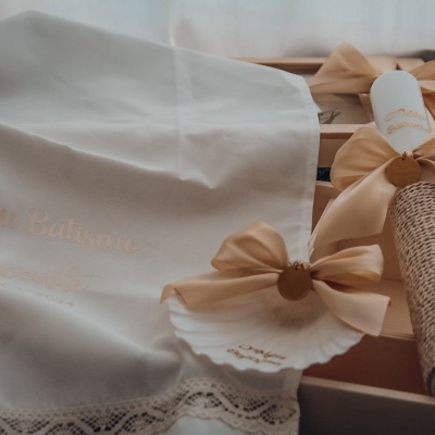 Toalha branca com renda e bordado dourado, rolo com tecido castanho claro e laço bege, caixa de madeira ao fundo