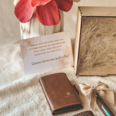 Conjunto de acessórios em pele castanha e cartão com texto, em fundo claro e flor vermelha ao fundo