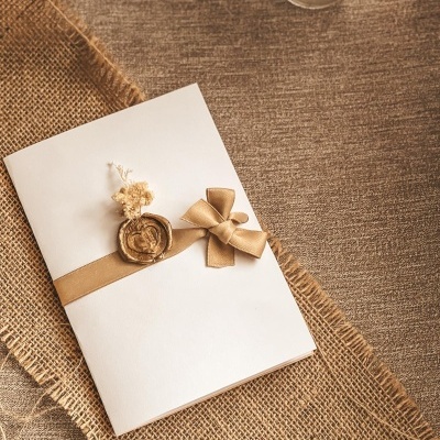 Convite branco com fita bege, laço e selo de cera, sobre tecido de juta texturado.