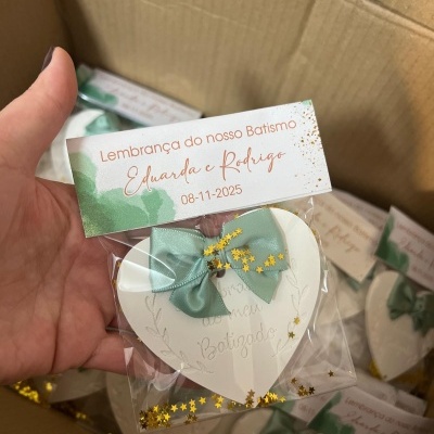Lembrança de batismo em placa branca de coração com laço verde e glitter dourado em embalagem transparente com etiqueta de batismo.