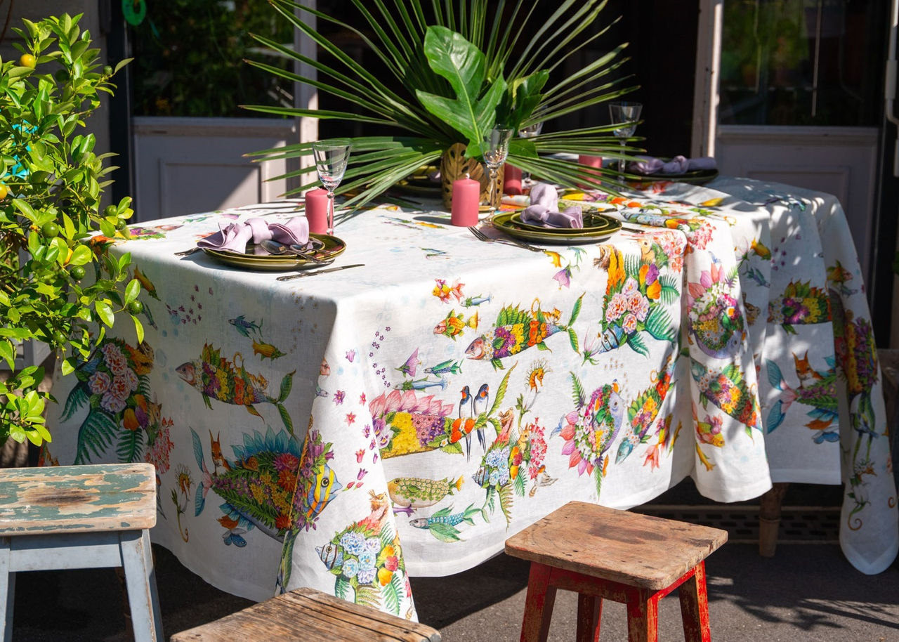 Toalha de Mesa 100% Linho PESCIFLORA- Tessitura Mesa exterior com toalha branca padrão pássaros e flores, pratos e guardanapos rosa, velas, bancos de madeira.