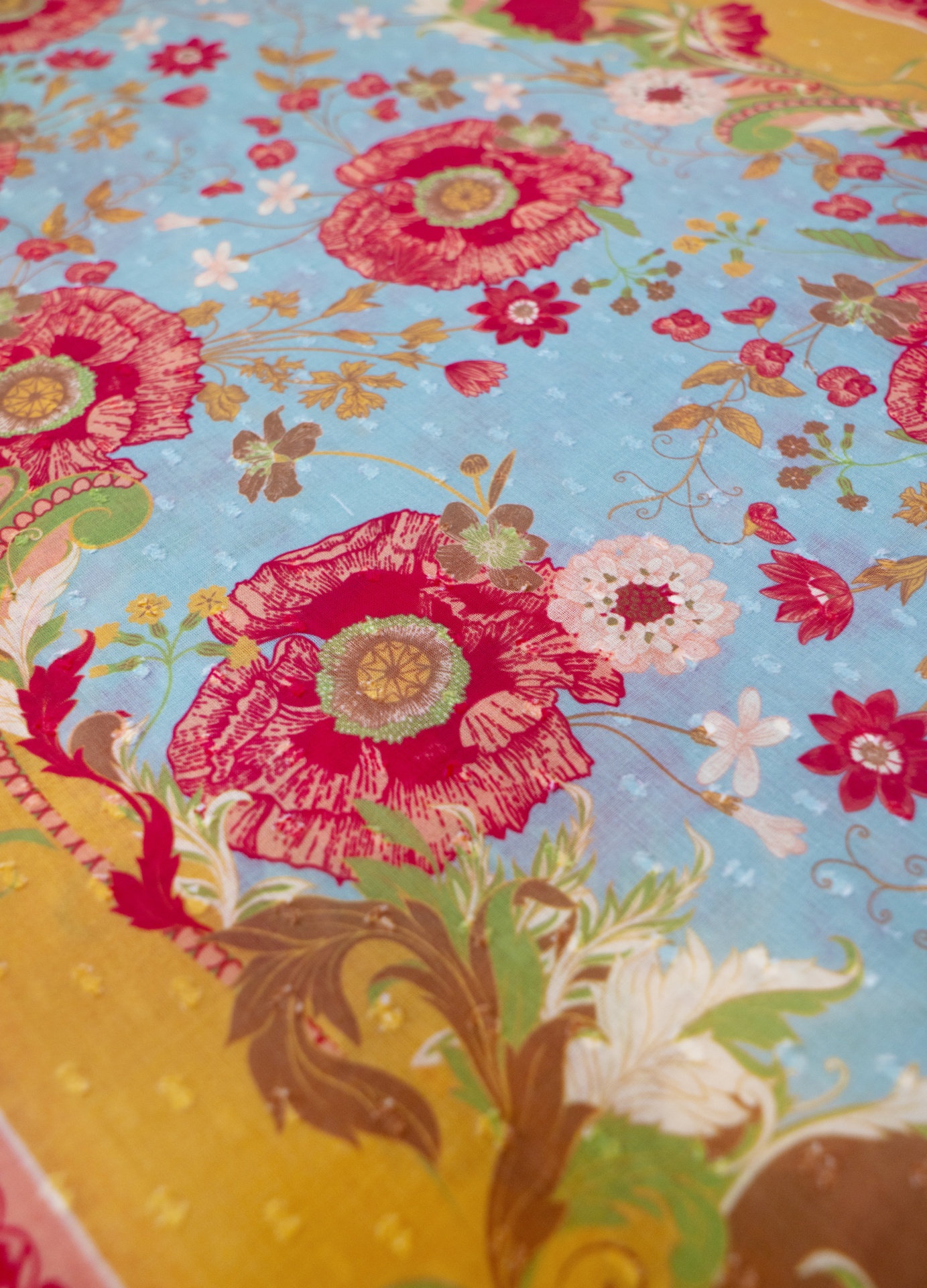 Echarpe Algodão Paisley Poppies- Powder Tecido com padrão floral colorido em fundo azul e detalhes decorativos.