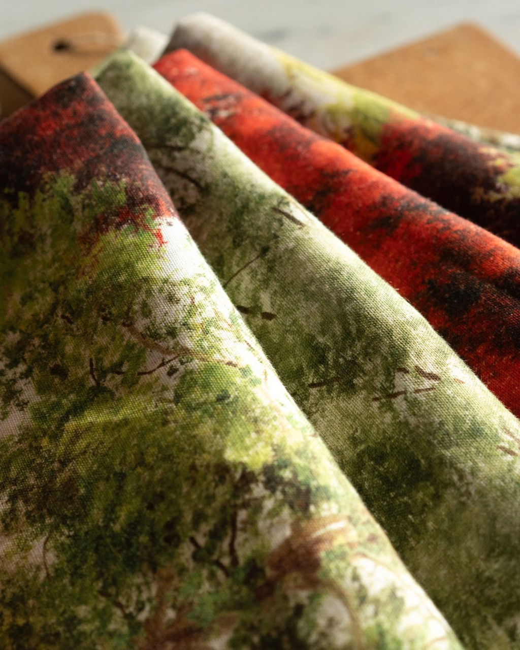 Toalha de Mesa 100% Linho GREESON- Tessitura Tecido estampado com desenhos de árvores em várias cores, dobrados sobre superfície de madeira.